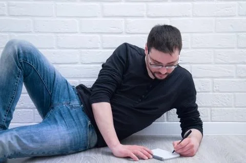 A young guy writes in a notebook, lying on a wooden floor against the backgro Stock Photos