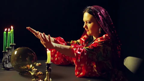 A young gypsy in a red dress at a table with candles looks into a glass ball and Stock Footage 99832845