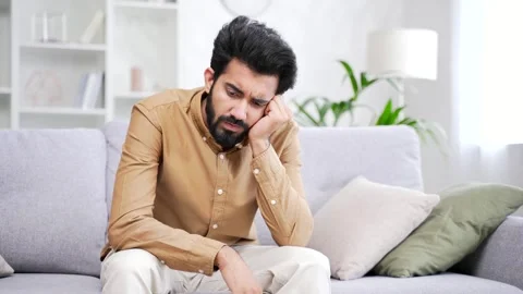 Young handsome bearded man feels depressed while sitting on sofa in living room  Stock Footage 252216103