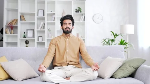 Young handsome bearded man meditating with eyes closed while sitting on sofa  Stock Footage 252215865