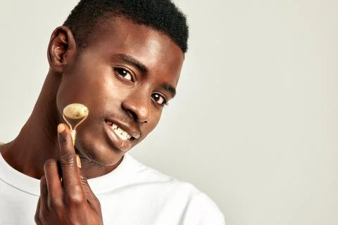 Young handsome black guy using jade de-puffing face roller to keep his skin soft Stock Photos