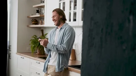 Young handsome blond man standing in the kitchen uses smartphone smiling and Stock Footage 239652276