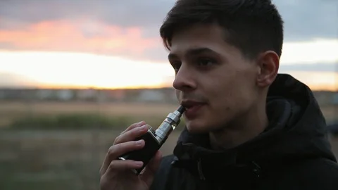 Young handsome boy with boxmod smoking e-cigarette at sunset outdoors Stock Footage 97537778