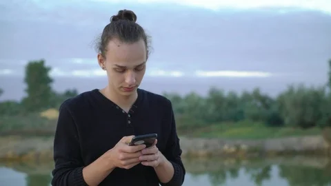 Young handsome boy using phone near the lake in the evening,4k Video stock 98501784