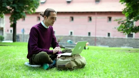 Young handsome brunette man using laptop sitting on grass outside in city. Urban Video stock 145756196