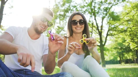 Young handsome couple sitting on the grass in the park and using hand spinners Vídeos de archivo 87911223