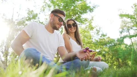 Young handsome couple sitting on the grass in the park and using hand spinners Stock Footage 87911235