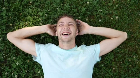 Young handsome guy lies on grass in summer park and laughs looking at camera Stock Footage 77443226