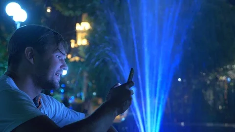 A young handsome guy sits in the evening next to a fountain with colored water Stock-Footage 93205637