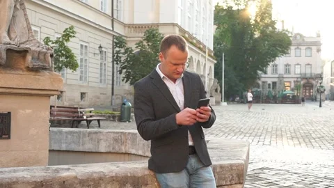 Young Handsome Guy using his Smartphone and Sitting on Old town Center fountain Stock Footage 164116364