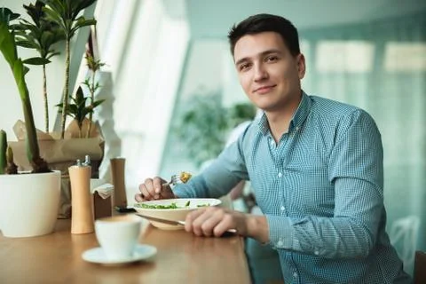 Young handsome happy man eats salad and drinks coffee for lunch during break at Stock Photos