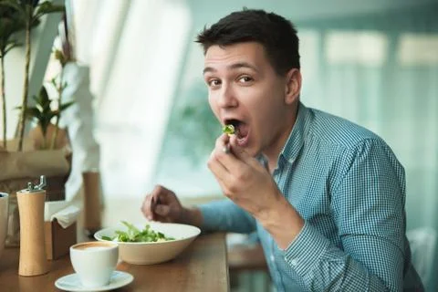 Young handsome hungty man eats salad and drinks coffee for lunch during break at Stock Photos