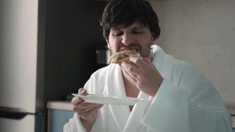 A young handsome man in a bathrobe is having breakfast, holding a croissant in Stock Footage 252660450