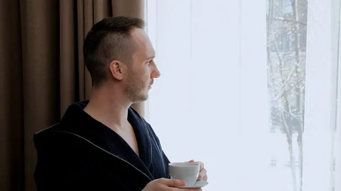 A young handsome man in a bathrobe stands at the window of the room and drinks Stock Footage 114316004