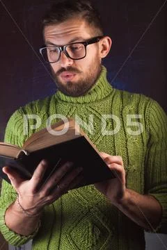 Young handsome man with a beard in green knitted sweater reading book Stock Photos