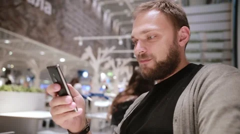 Young handsome man with a beard sits in a cafe, uses his smartphone, smiles and Stock Footage 67947735