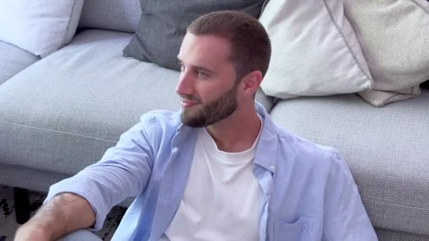 Young handsome man with a beard sitting on the floor leaning against a sofa Vídeos de archivo 345113671