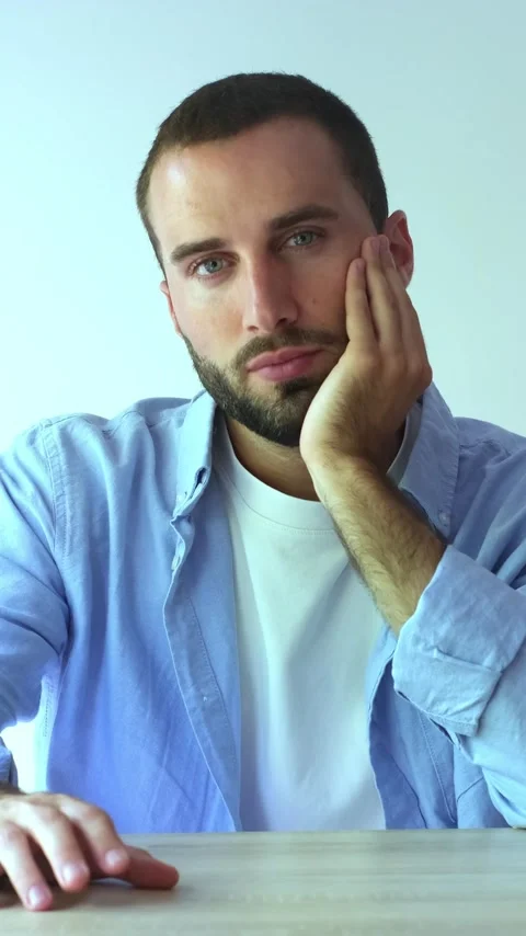 Young handsome man with a beard sitting at a table, looking sad and tired Stock Footage 345123875