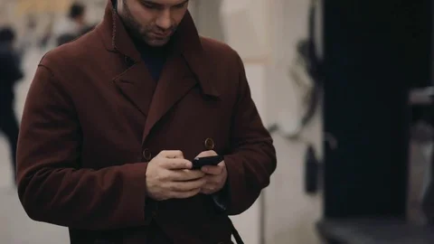 Young handsome man in a coat standing in the city center and using phone Video stock 74806501