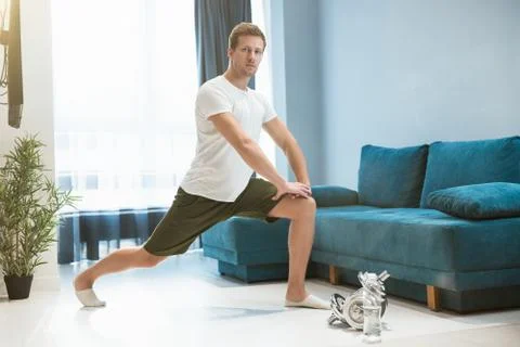 Young handsome man doing lunges during stretching before workout at home looking Stock Photos