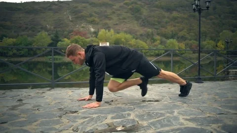 A young handsome man doing mounting climber exercise on a terrace near the pond Stock Footage 70345067