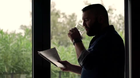 Young, handsome man drinking water and reading newspaper by the window HD Stock Footage 57361303