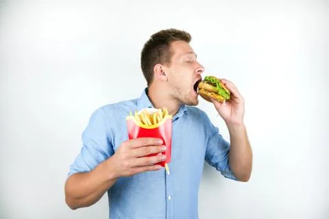 Young handsome man eats cheeseburger and holds french chips from fast food Stock Photos