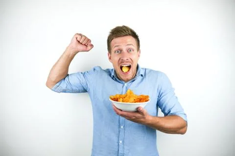 Young handsome man eats chips holding his fist up happily on isolated white Stock Photos