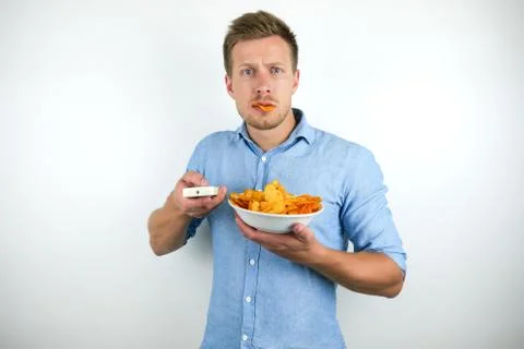 Young handsome man eats chips holds remote control on isolated white background Stock Photos