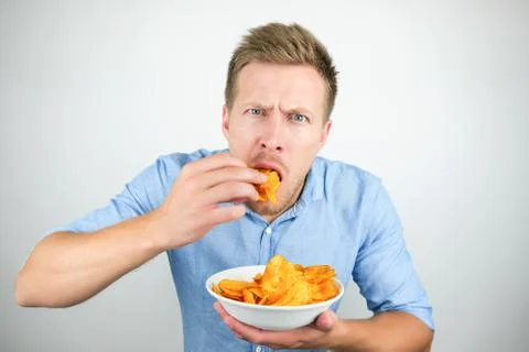Young handsome man eats chips with paprika from plate looking hungry on isolated Stock Photos