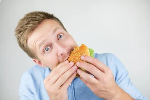 Young handsome man eats fresh tasty burger from fast food restaurant looks Stock Photos
