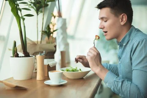 Young handsome man eats salad and drinks coffee for lunch during break at cafe Stock Photos
