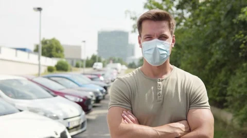 A young handsome man in a face mask looks at the camera in a parking lot in an 스톡 동영상 145192075
