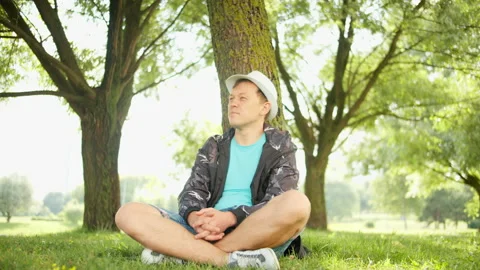 Young handsome man in a hat sits under a tree in a city park, camera movement Stock Footage 138998182