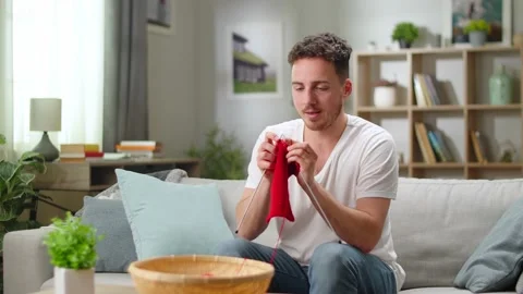 Young handsome man knitting red scarf with knitting needles while sitting on Video stock 144179934