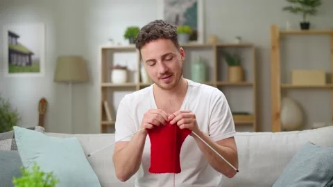 Young handsome man knitting a red scarf with needles for the new year sitting in Video stock 144179968