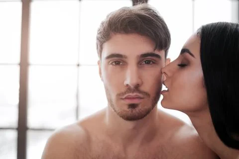 Young handsome man look on camera. Beautiful woman kiss him. Window is behind Stock Photos