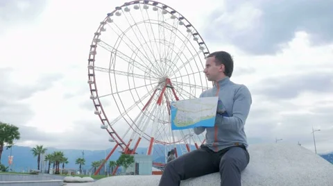Young handsome man looking for landmarks on a paper map nearby ferris wheel Stock Footage 67611823