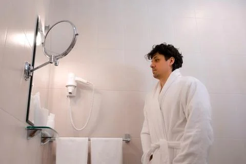 Young handsome man puts on a white robe in the bathroom looking at mirror. Stock Photos