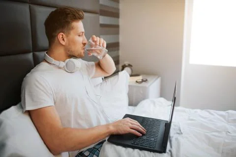 Young handsome man sit in bed this morning. He drink water. Male model hold Stock Photos