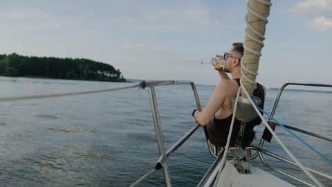 A young handsome man sits on the bow of a sailing boat and drinks wine in summer Stock Footage 235993913