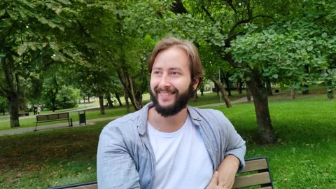 A young handsome man is sitting on a bench in a city public park. A cute guy Stock Footage 227932619