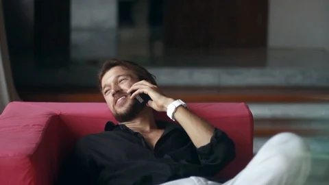 Young handsome man is sitting on the couch, talking on the phone and using a Stock-Footage 85525946