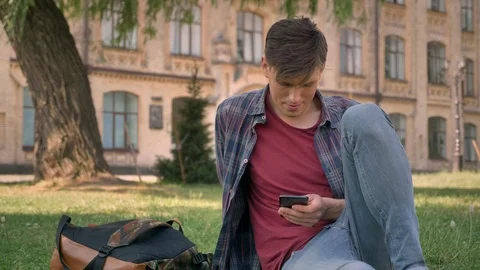 Young handsome man is sitting on grass in park, watching pages in Internet on Video stock 91728080