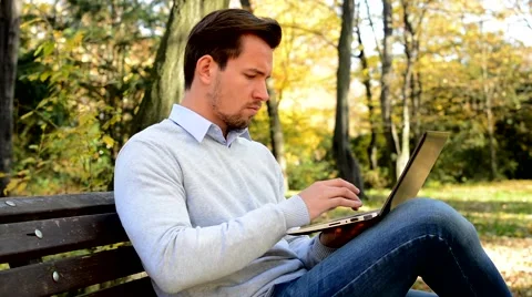 Young handsome man sitts on bench in a park and works with a laptop Stock-Footage 59725823