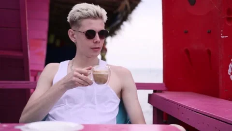 Young handsome man in sunglasses drinking latte in summer cafe on the seashore Stock Footage 233587241