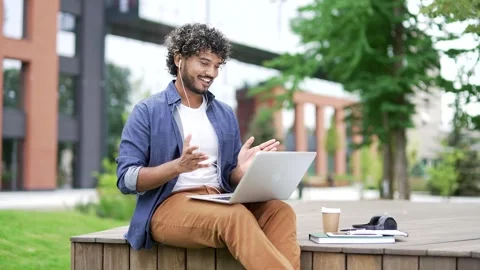 Young handsome man talking on a video call using laptop sitting on a bench near  Stock Footage 302472397