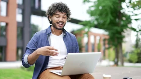 Young handsome man talking on a video call using laptop sitting on a bench near  Stock Footage 317710738