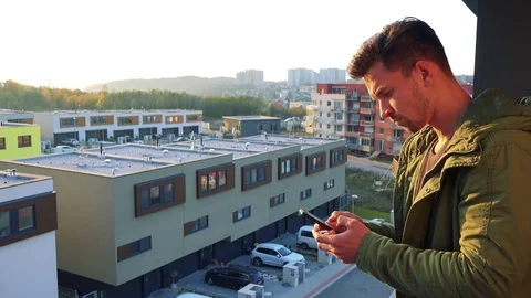 A young, handsome man types on his smartphone on a balcony, eventually looks out 库存影片 71175191
