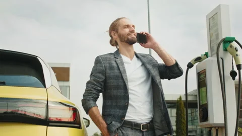 Young handsome man use phone while his electro car charging at station. Electric Stock Footage 235361417